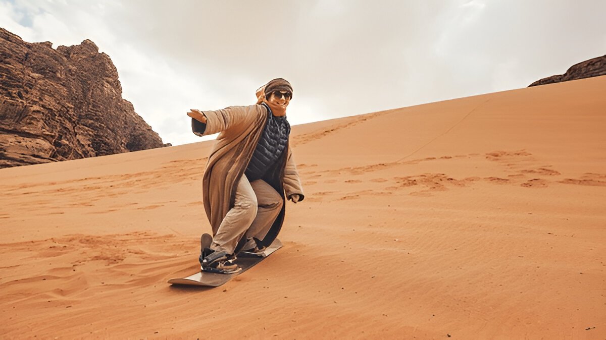 Exciting scene of sandboarding in the Dubai desert with a person surfing down a steep sand dune, kicking up sand under a bright blue sky. @exploredesertsafari.com