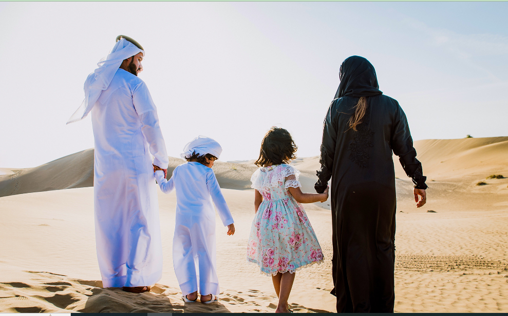 Vibrant scene in the Dubai desert with guests dressed in traditional Arabic costumes, including abayas and kanduras, at a desert camp with sand dunes in the background." @exploredesertsafari.com