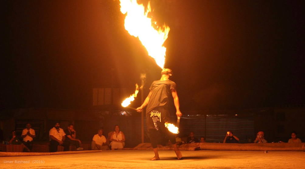 Exhilarating fire show in the Dubai desert with a performer spinning flames under a starry night sky, illuminating the sand dunes and audience." @exploredesertsafari.com