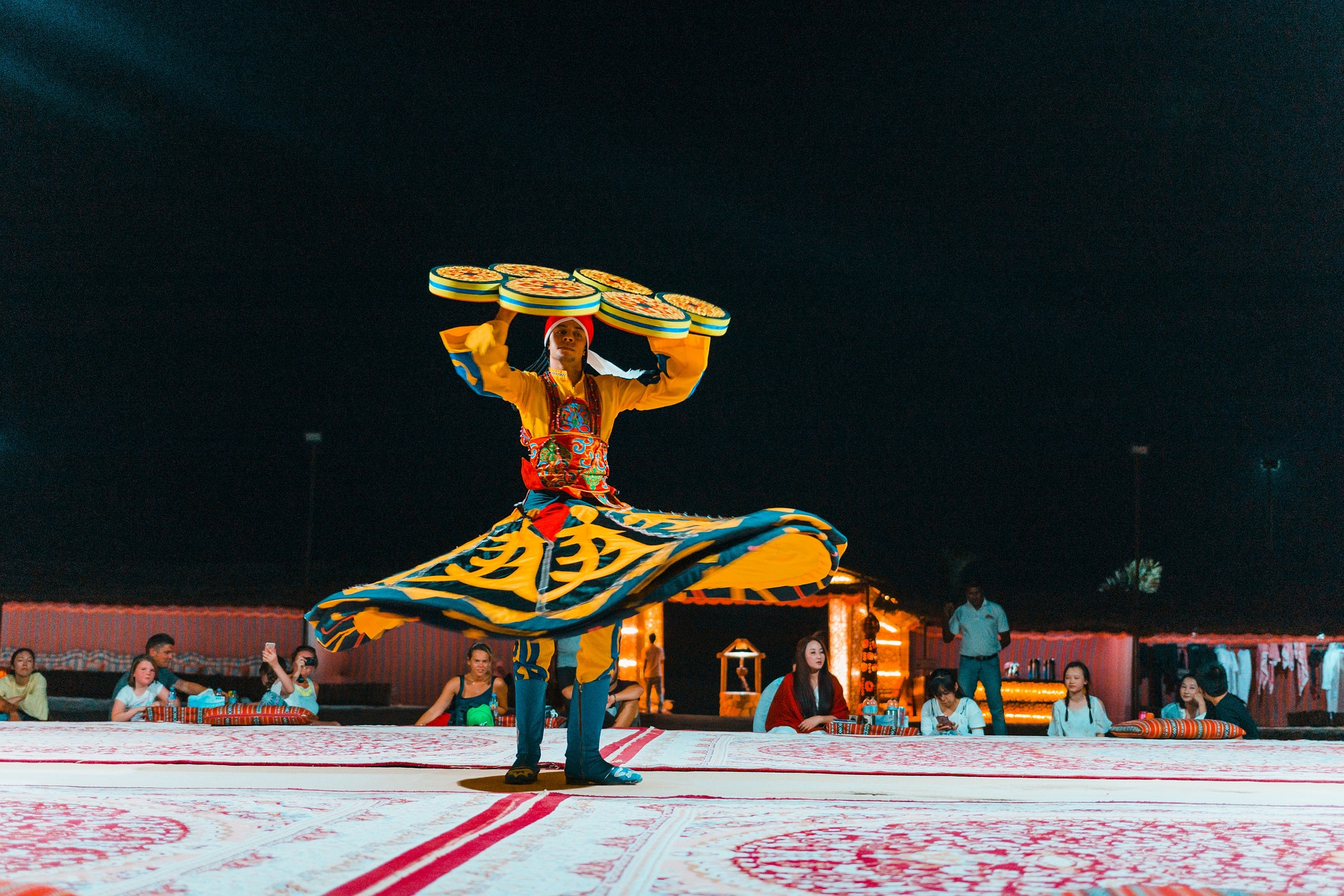 Vibrant Tanoura show in the Dubai desert with a dancer spinning in brightly colored, multi-layered skirts, and an audience seated under a starry sky." @exploredesertsafari.com